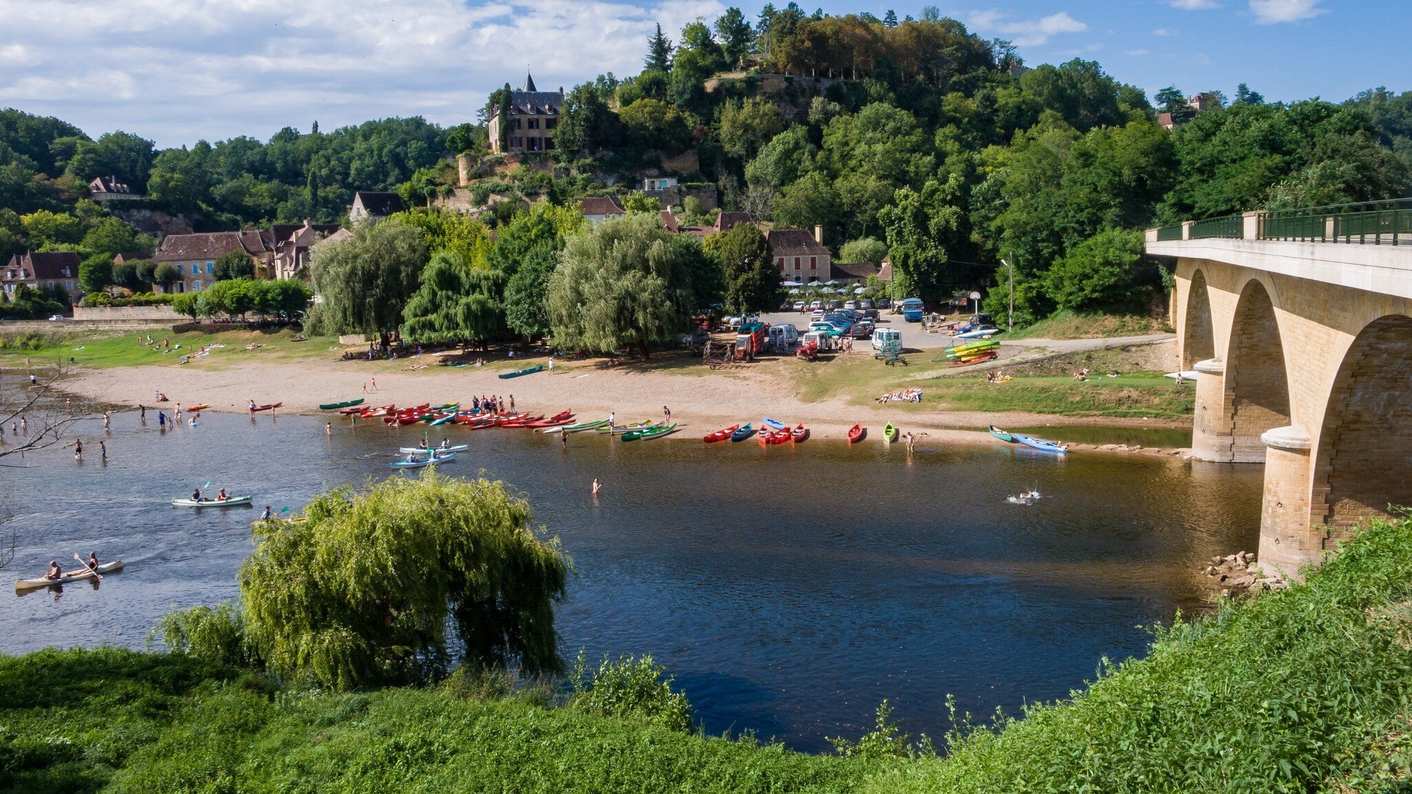 Canoes and swimmers on the Dordogne River beside a stone bridge below the hillside village of Limeuil, France