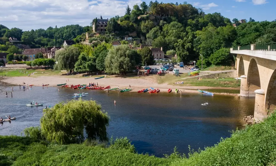 Canoes and swimmers on the Dordogne River beside a stone bridge below the hillside village of Limeuil, France