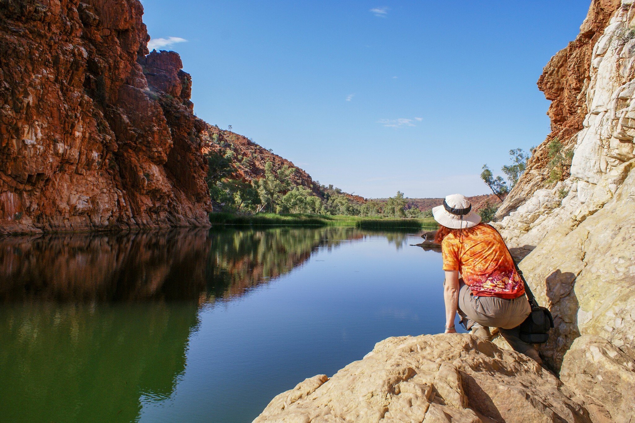 Woman with hat kneeling on a rock beside the waterhole at Glen Helen Gorge, West MacDonnell Ranges, Australia.