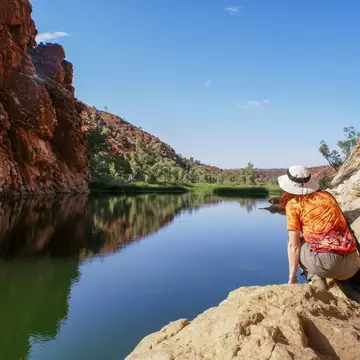 Woman with hat kneeling on a rock beside the waterhole at Glen Helen Gorge, West MacDonnell Ranges, Australia.