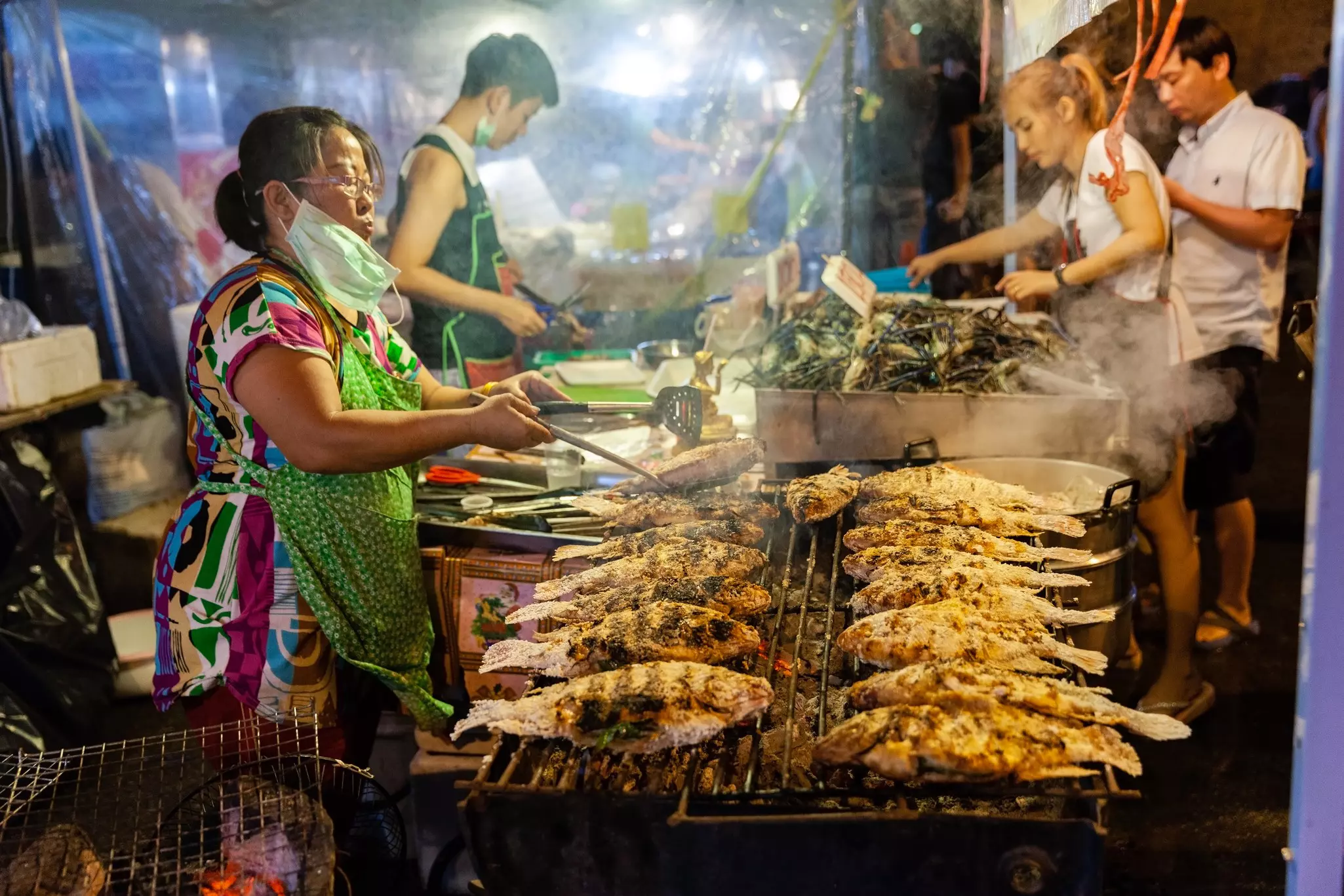A vendor cooks fish on a griddle in a smokey street food market.