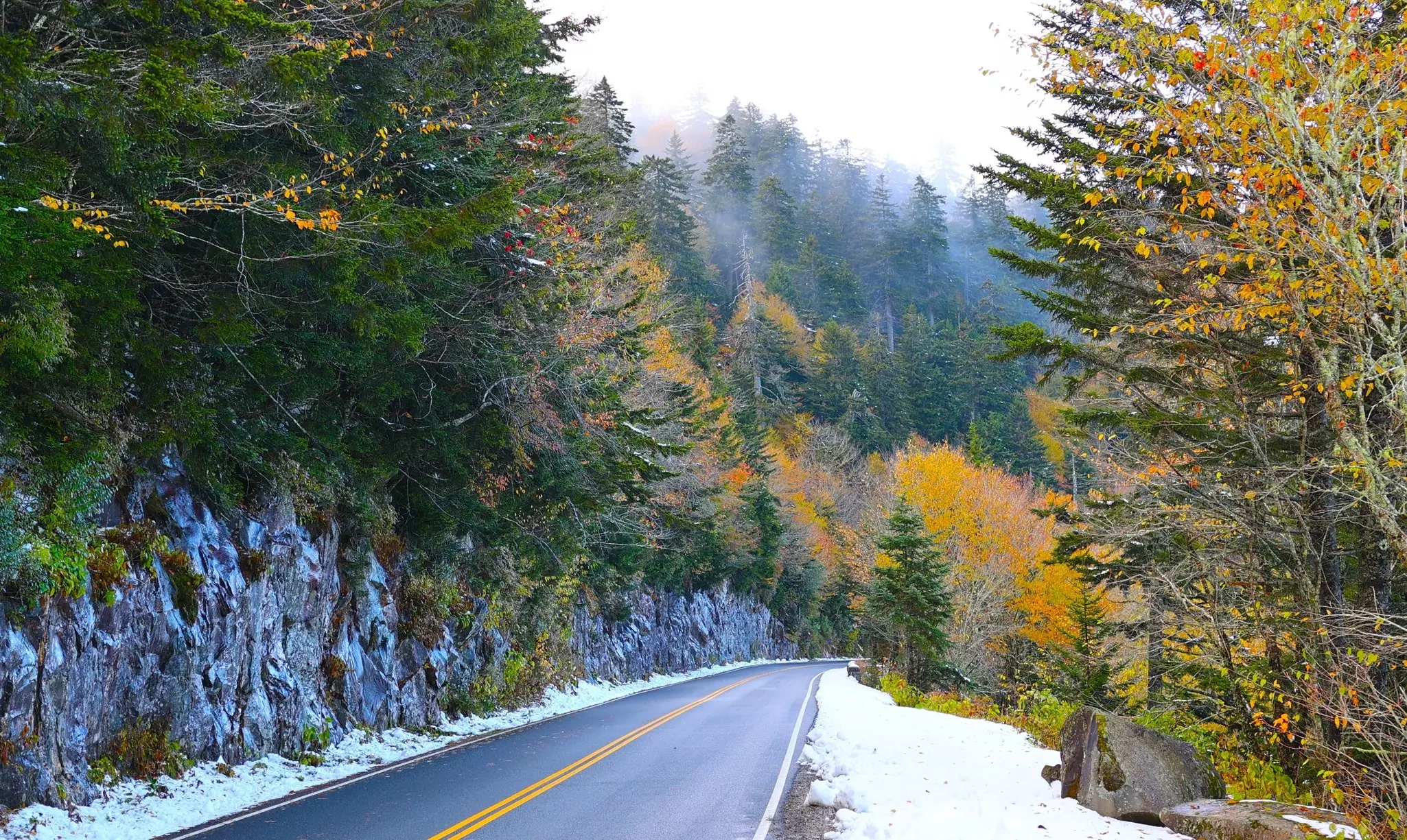 Patches of snow appear next to a blacktop roadway; trees have autumn foliage.