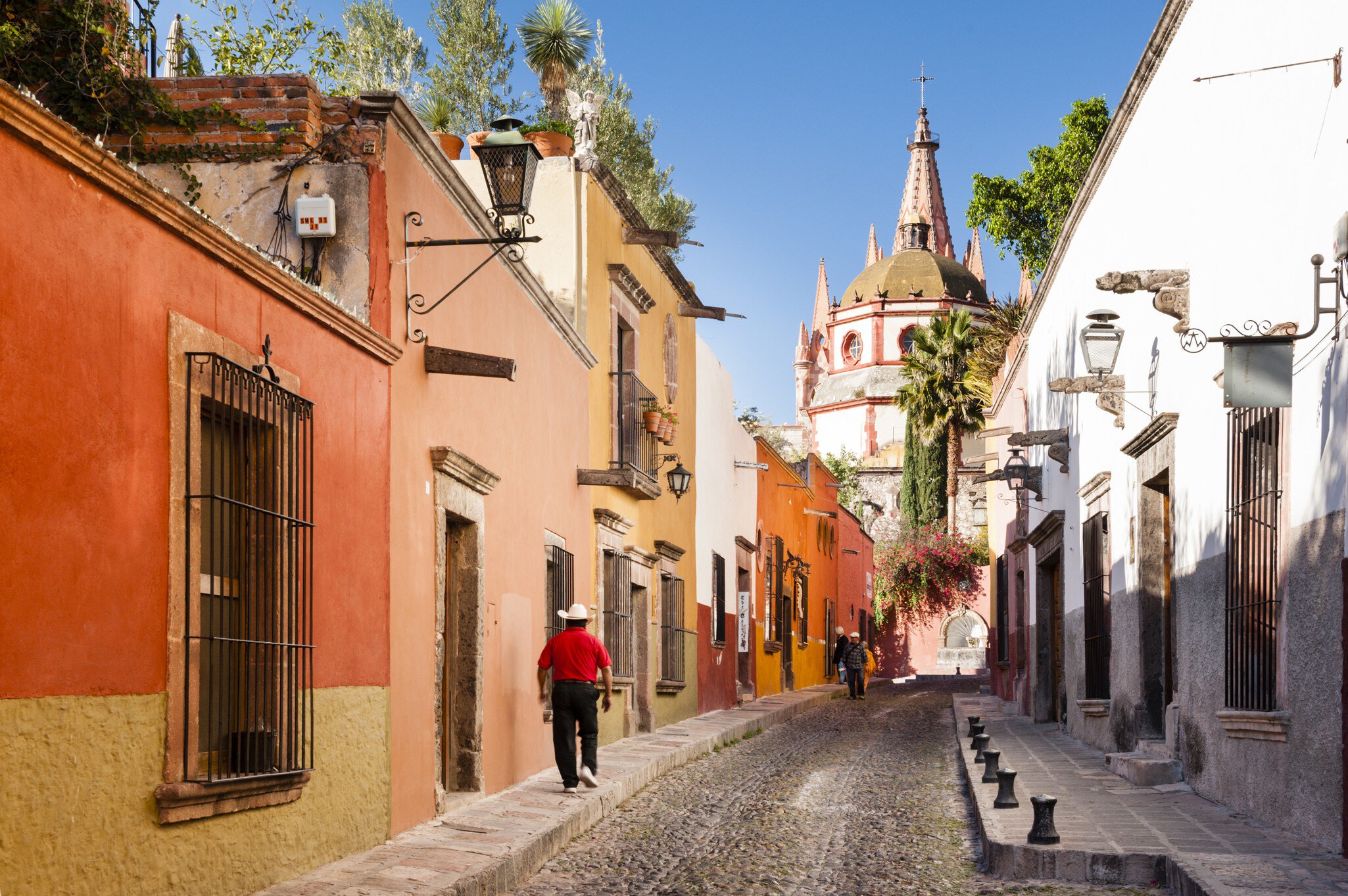 A man in a cowboy had walks by brightly painted buildings on a narrow cobblestone street. A church dome is seen in the distance.