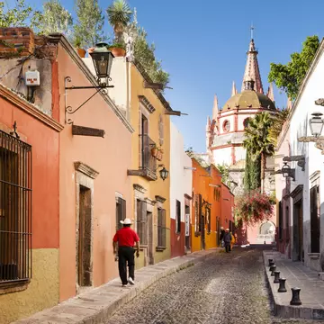 A man in a cowboy had walks by brightly painted buildings on a narrow cobblestone street. A church dome is seen in the distance.