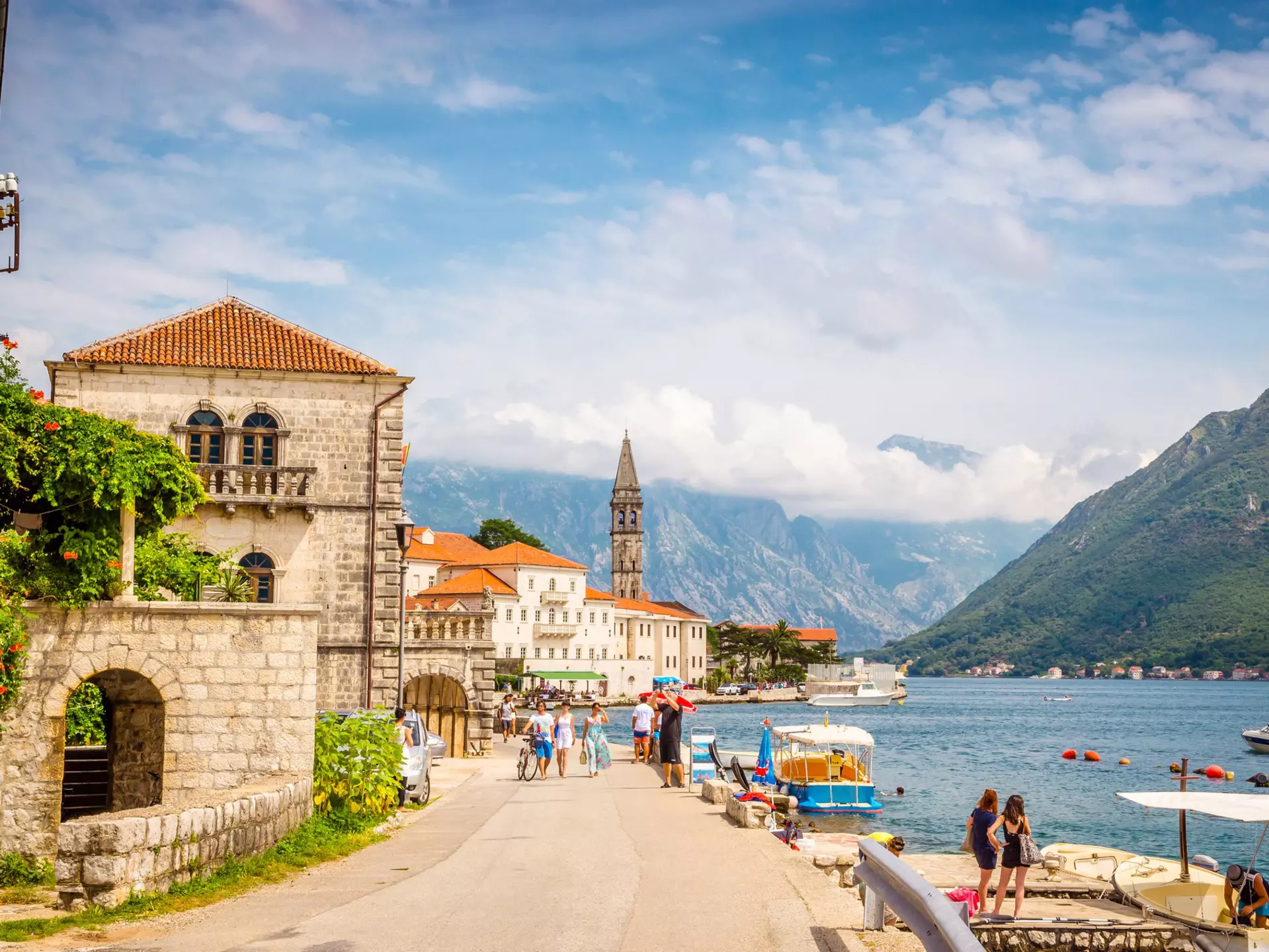 People walking on a shoreline promenade amid low-profile buildings with a mountain backdrop in Perast, Montenegro