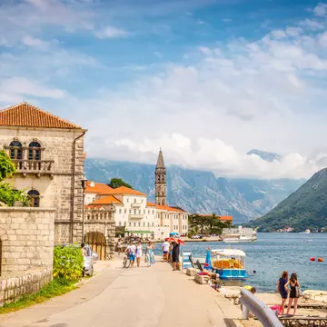 People walking on a shoreline promenade amid low-profile buildings with a mountain backdrop in Perast, Montenegro