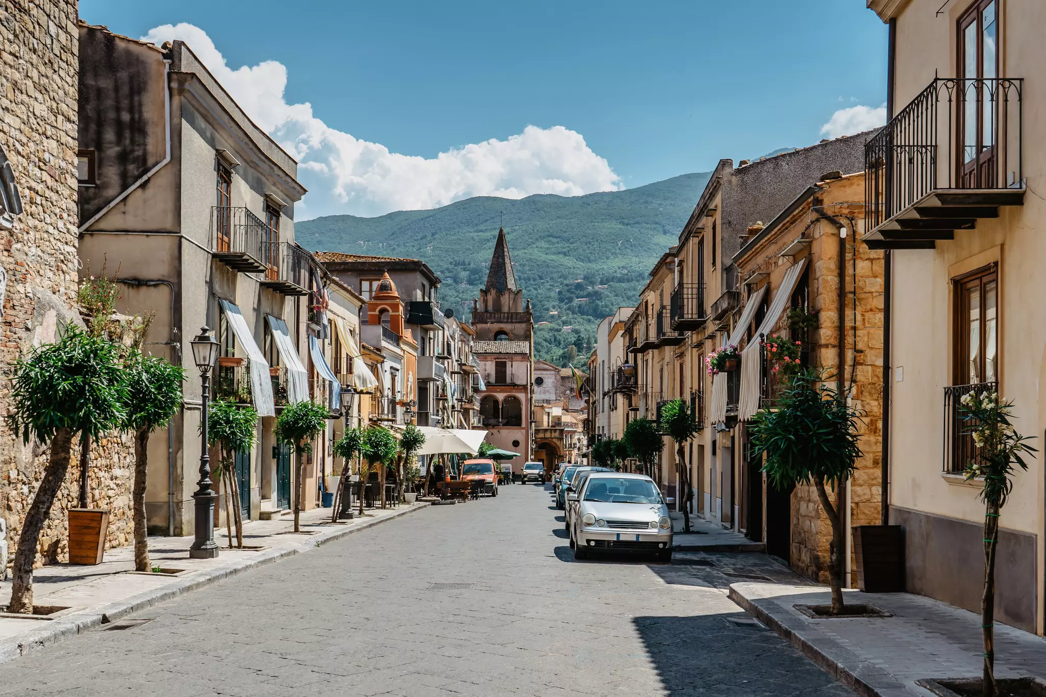 A medieval street with cars parked down one side of it. Hills rise above a church in the distance.