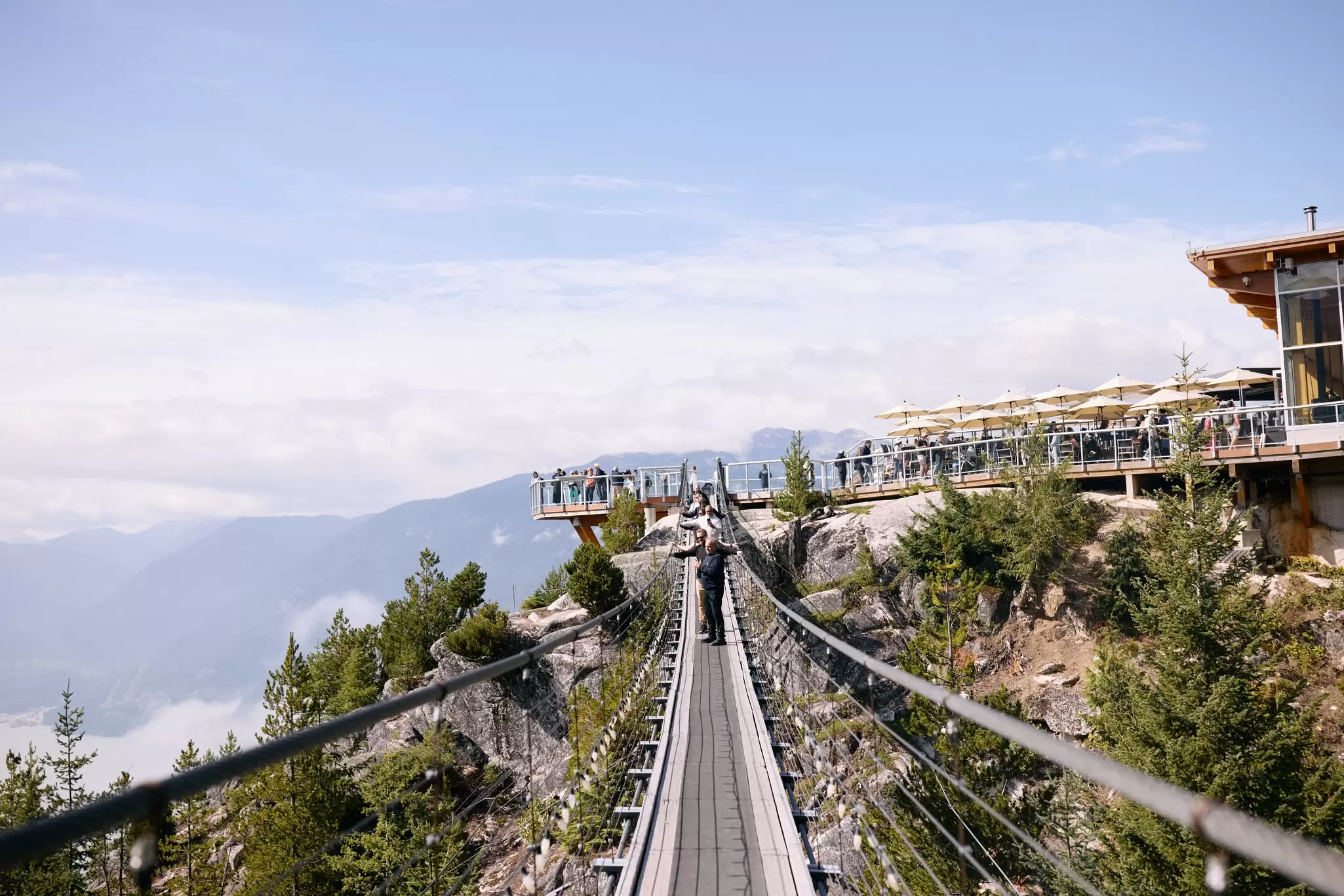 View from one end of a long hanging bridge as tourists are crossing it from the other direction. Mountains in the background, and a building with a lookout with umbrellas is on the other end of the bridge.