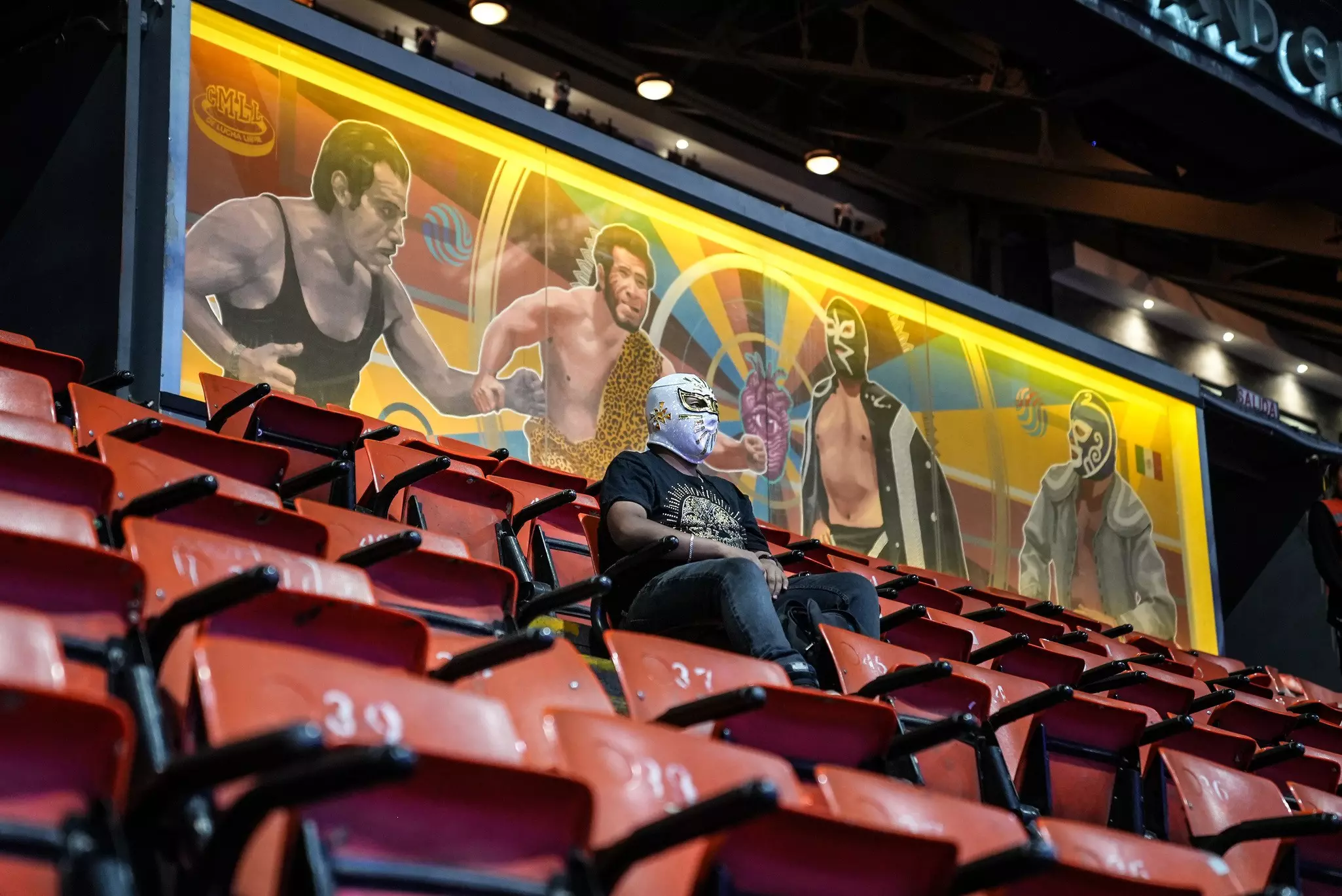 A person sits below a mural in a stadium wearing a lucha libre mask; he is surrounded by empty seats.