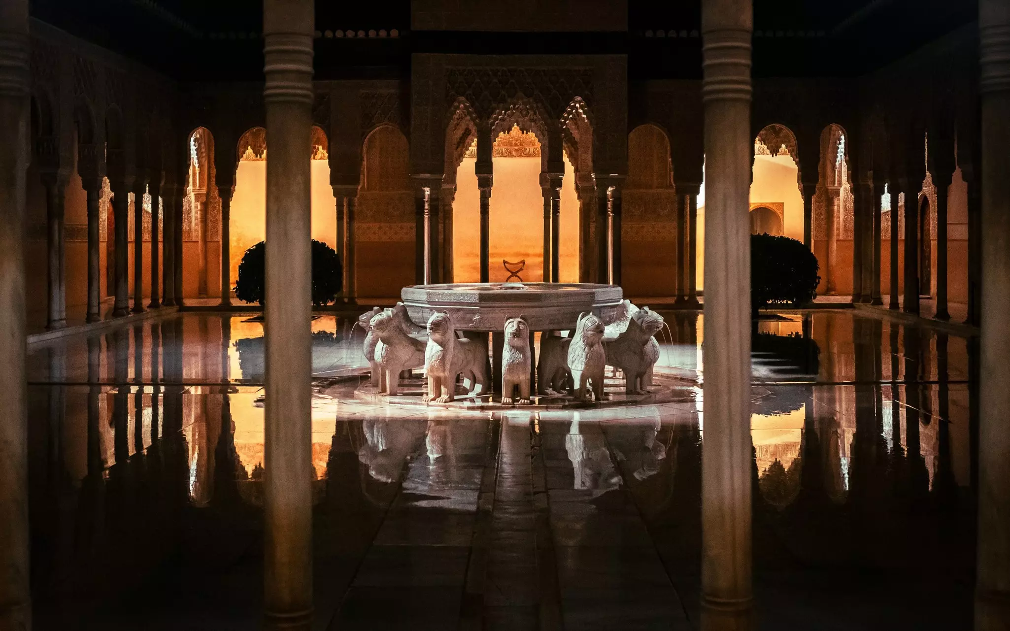 A fountain encircled by sculptures of lions is in the center of a courtyard; the stone is wet, and the fountain and surrounding columns and archways are reflected on the water's surface.