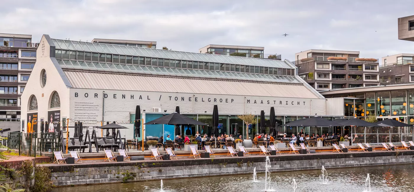 A modern industrial building turned theater space with large glass windows overlooking a lake. People are seated at an outdoor terrace in front of the building.