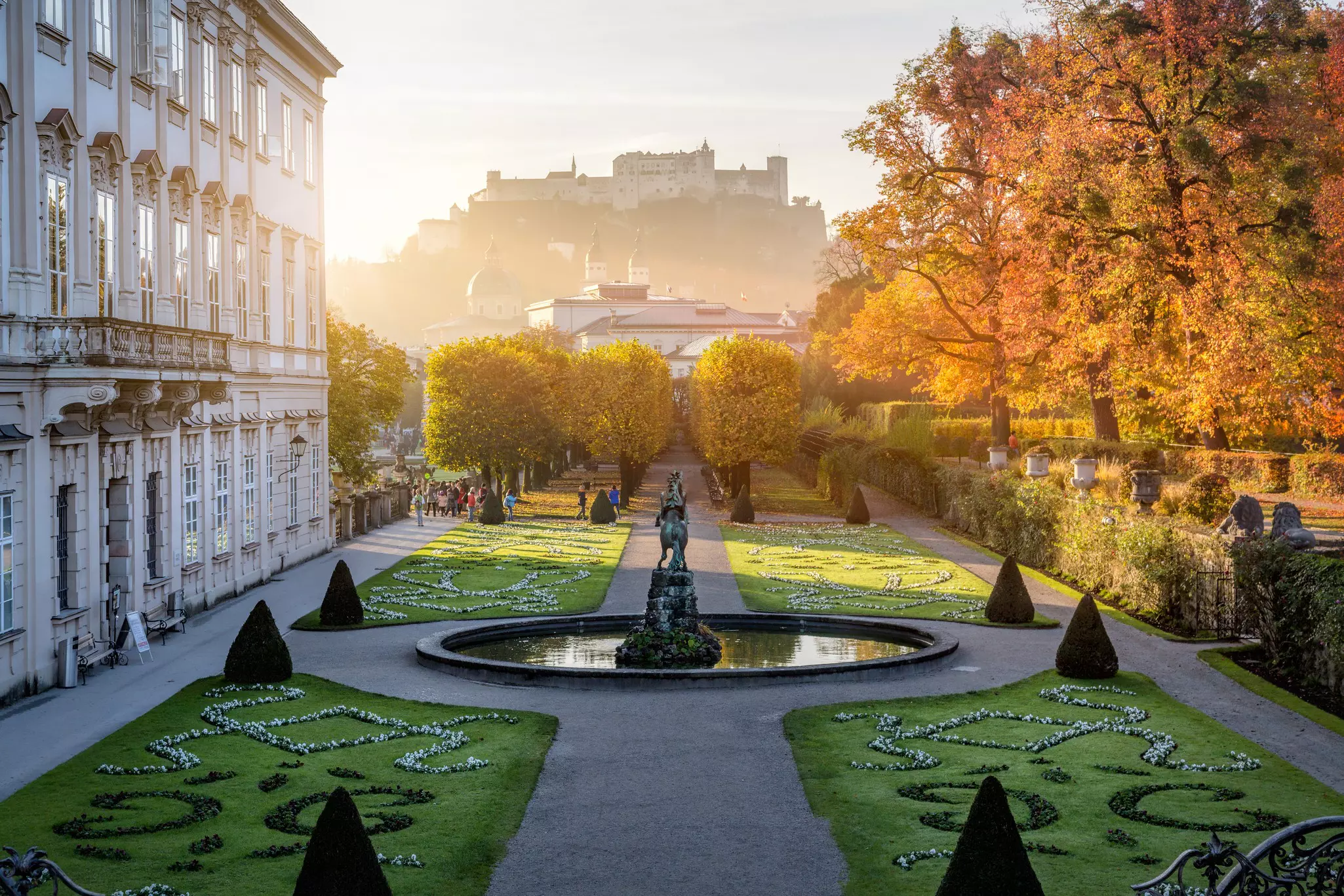 Garden with designs in the grass and cone-trimmed evergreens with historic building to the left, fountain with bronze sculpture in the center, autumn-leaved trees to the right, and historic buildings on a hill in the far distance as the sun rises.
