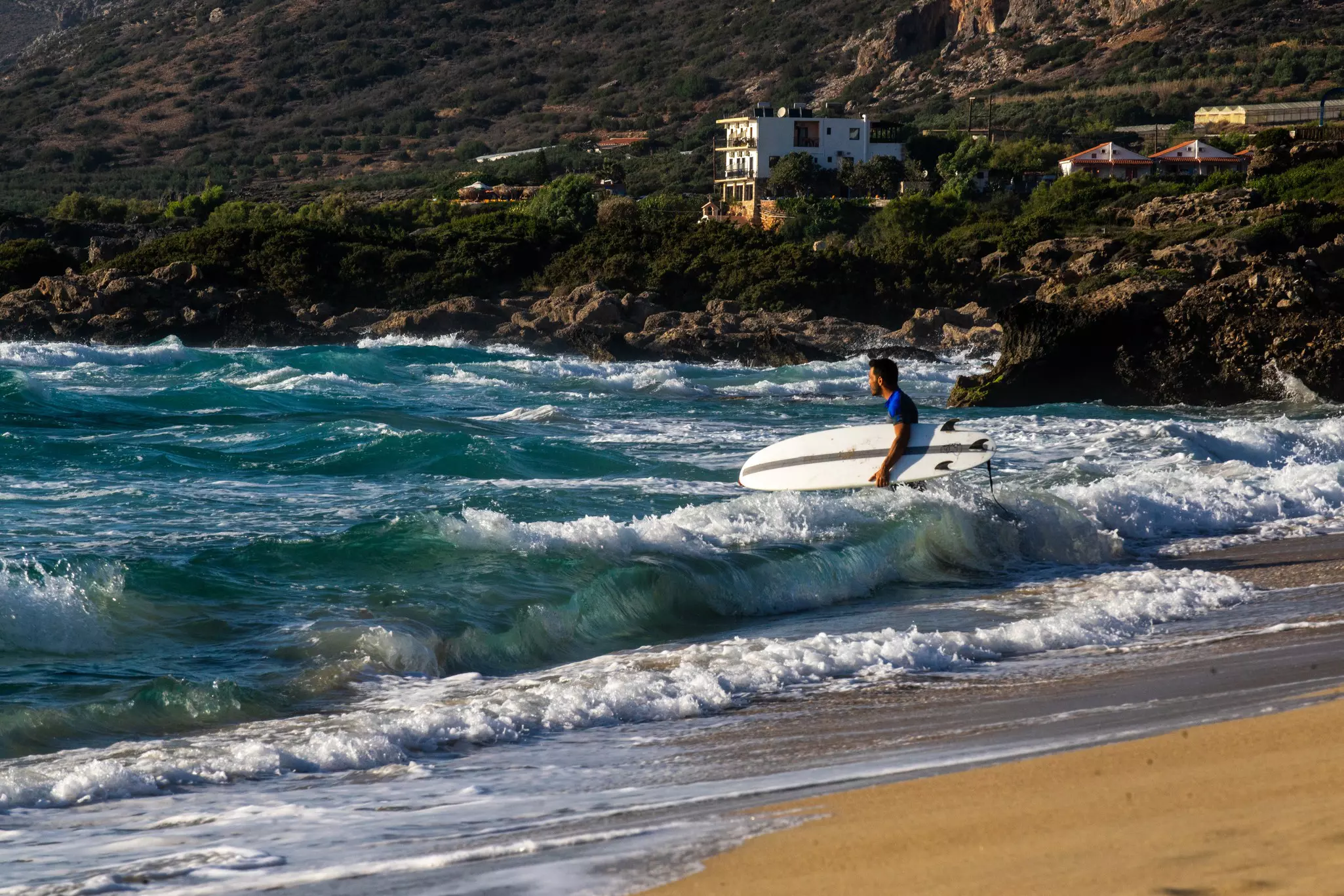 A surfer holding his board wades into white-capped waves at beach.