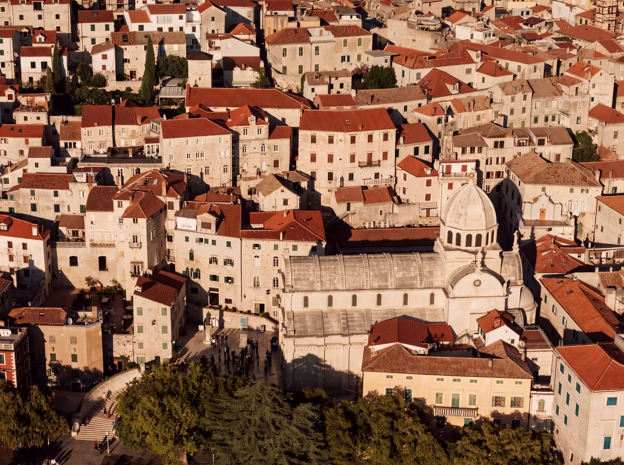 St. James cathedral in Sibenik, Croatia is white with steps leading to its door that wrap around some trees in front of the building.