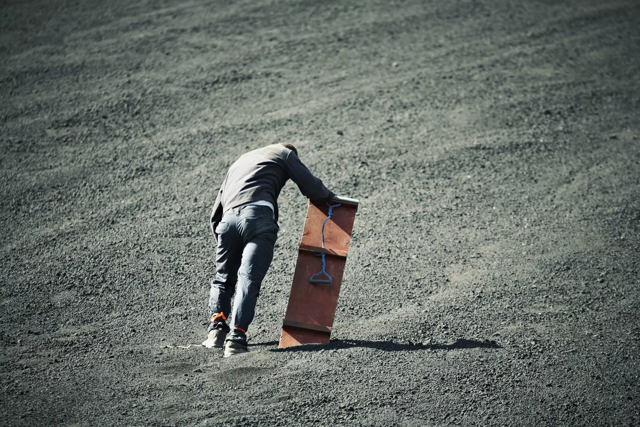 A tired man holding a sandboard on a dusty volcanic landscape.