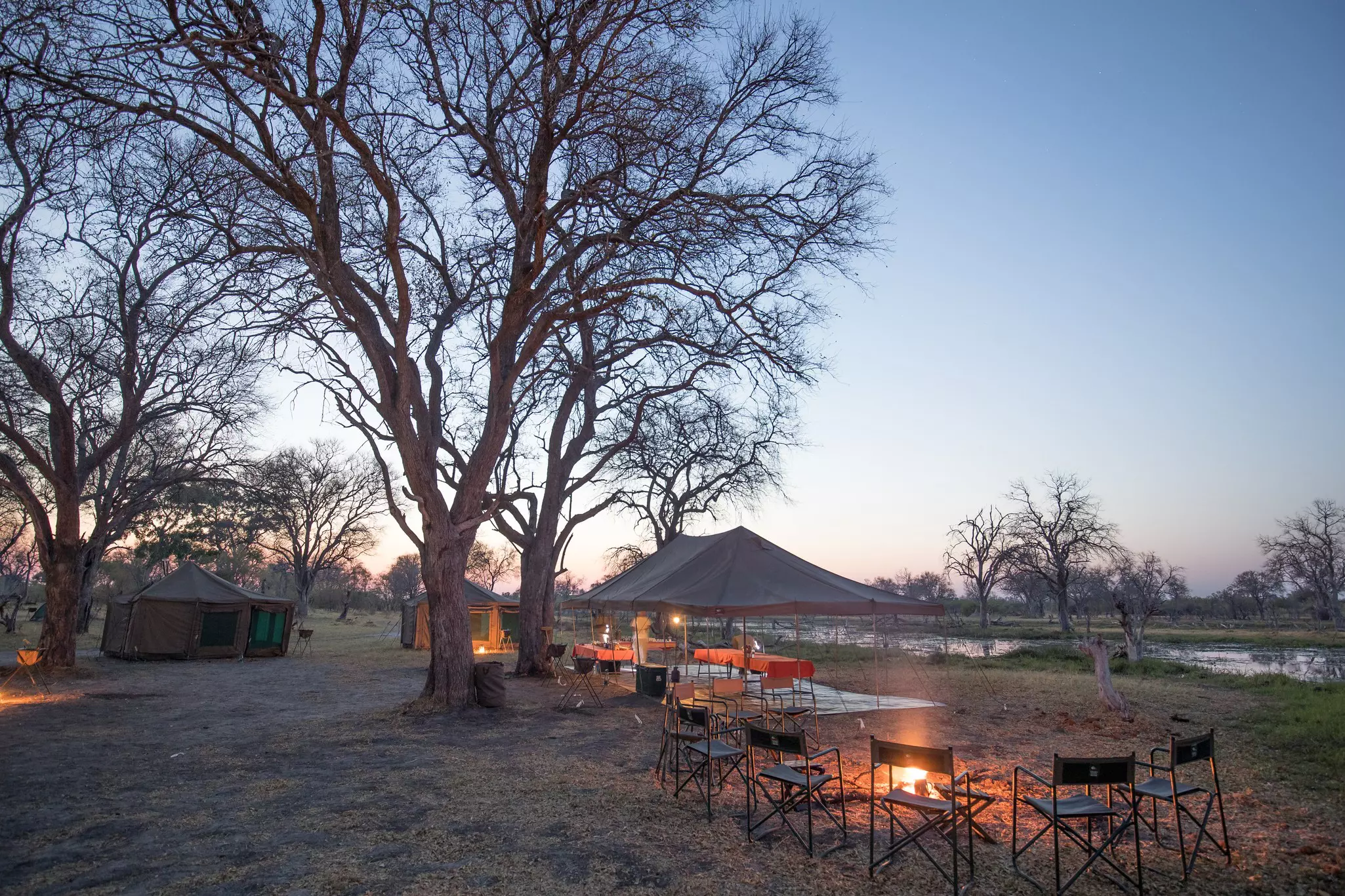 Camp chairs around a fire and a large canopy set up at dusk at a campsite near a river.