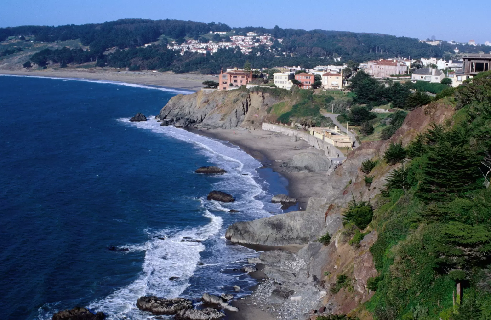 Rugged coastline where waves break on a sandy cove below steep cliffs topped by hillside houses, under a clear blue sky.