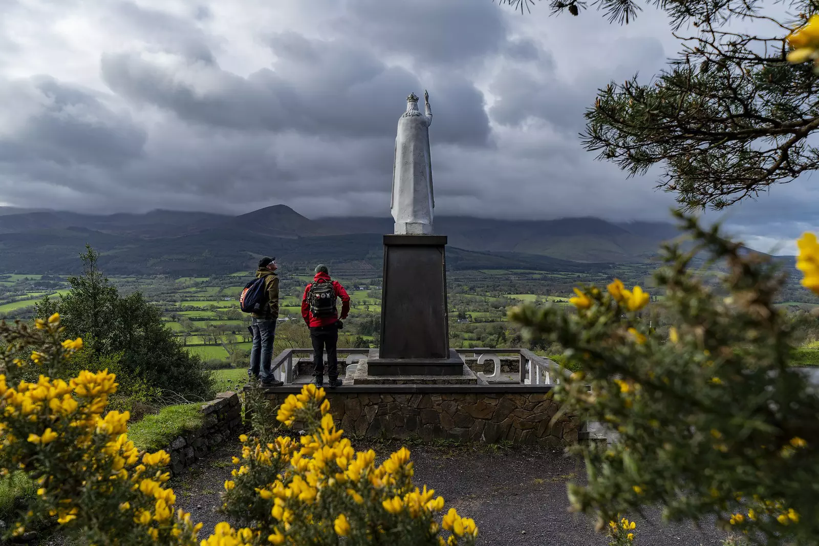 Two hikers stand beside a religious statue that overlooks a mountain vale on a spring day