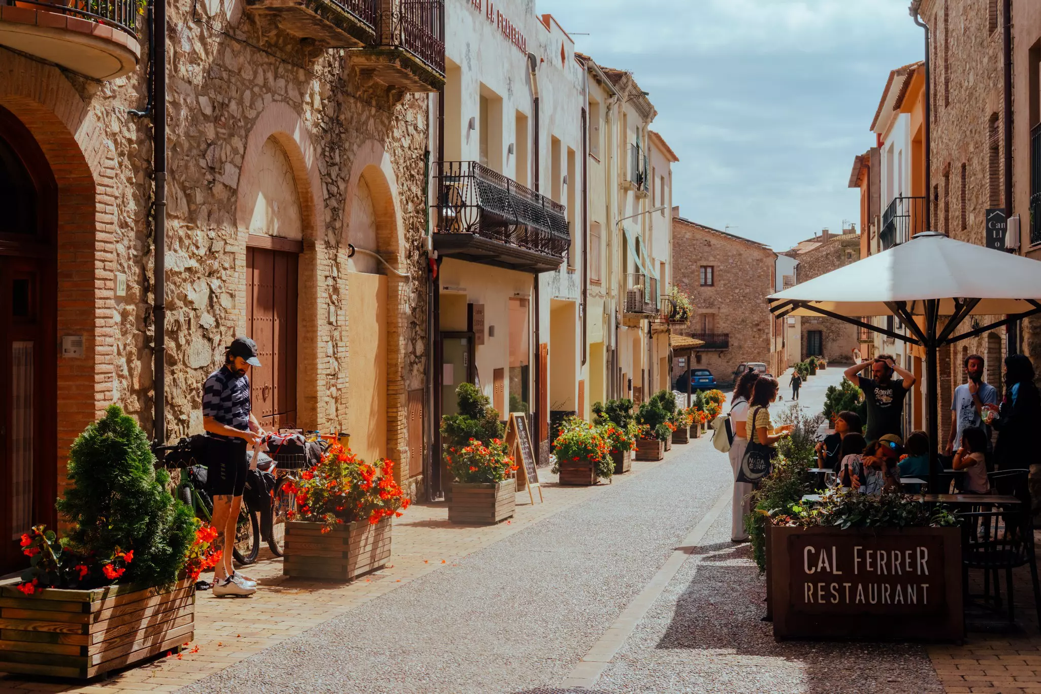 A sunny street in a small town in Spain; a person stands near a bike to one side, and a cafe has outdoor tables in the shade of an umbrella on the other side.