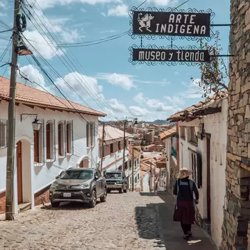 A wrought-iron sign reading ARTE INDIGENA, museo y tienda hanging over a steep cobbled street of whitewashed and stone buildings, a woman in a straw hat walking down it.