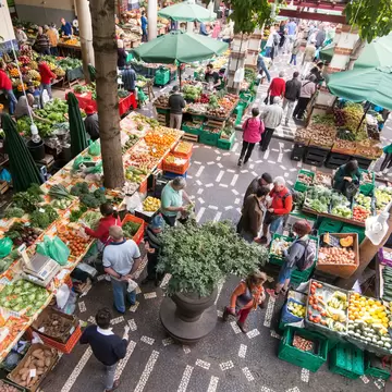 Looking down into a covered market courtyard of fruit and vegetable stalls under green parasols, shoppers moving between them on patterned stone paving.