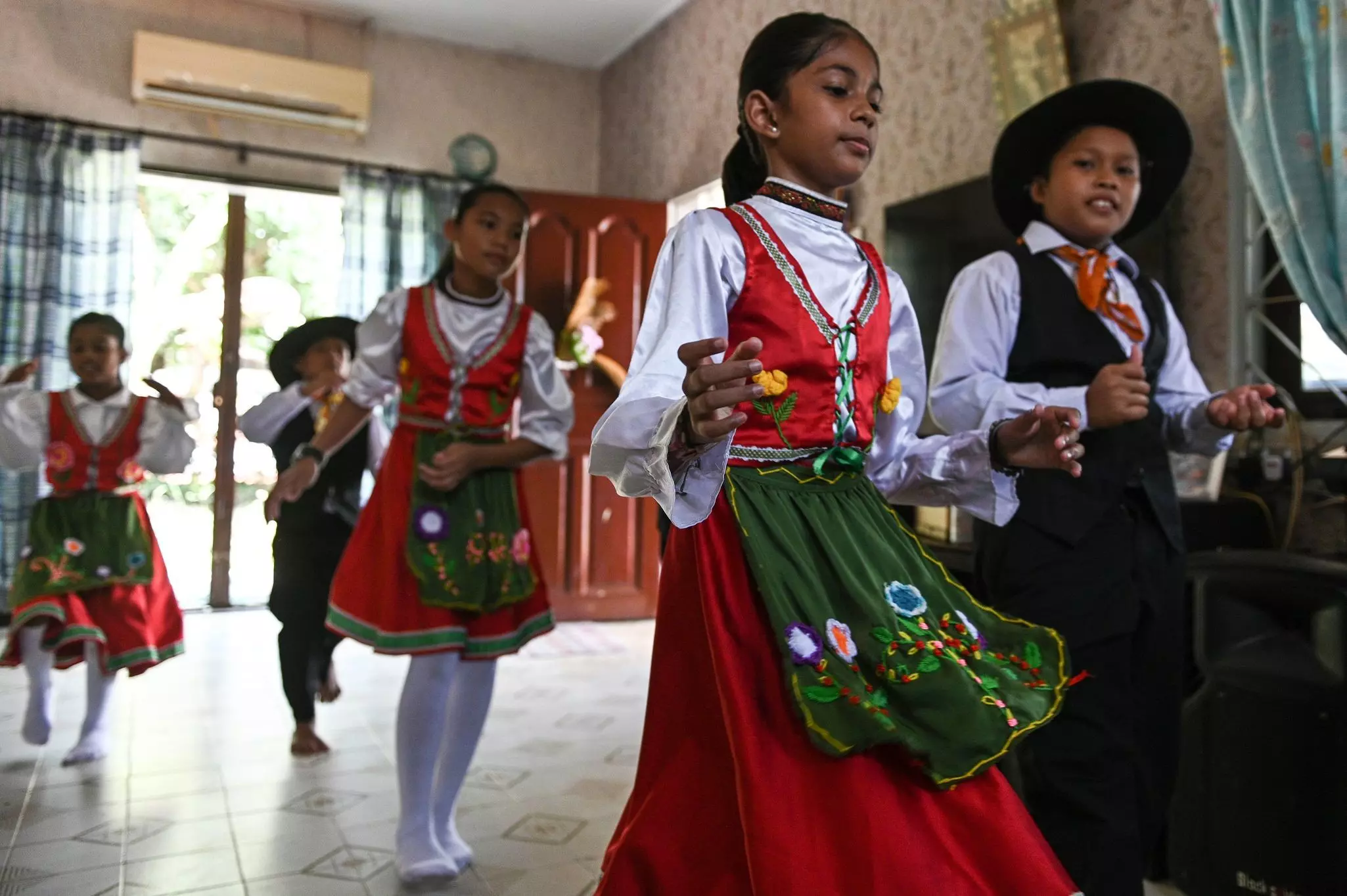 Children in traditional costumes dance in a classroom.