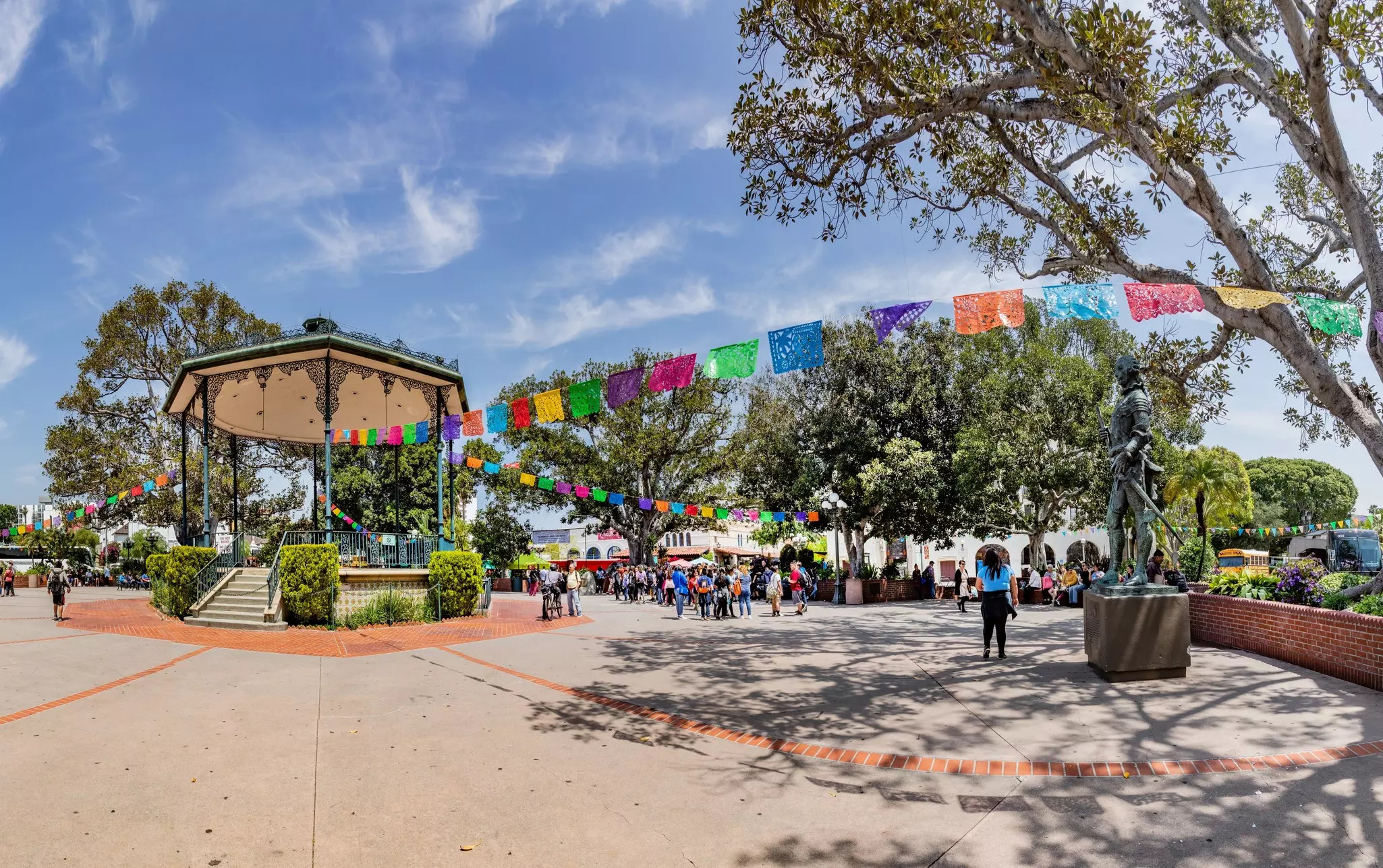 A large city square with a central bandstand. People shop at the nearby market under colorful decorations.