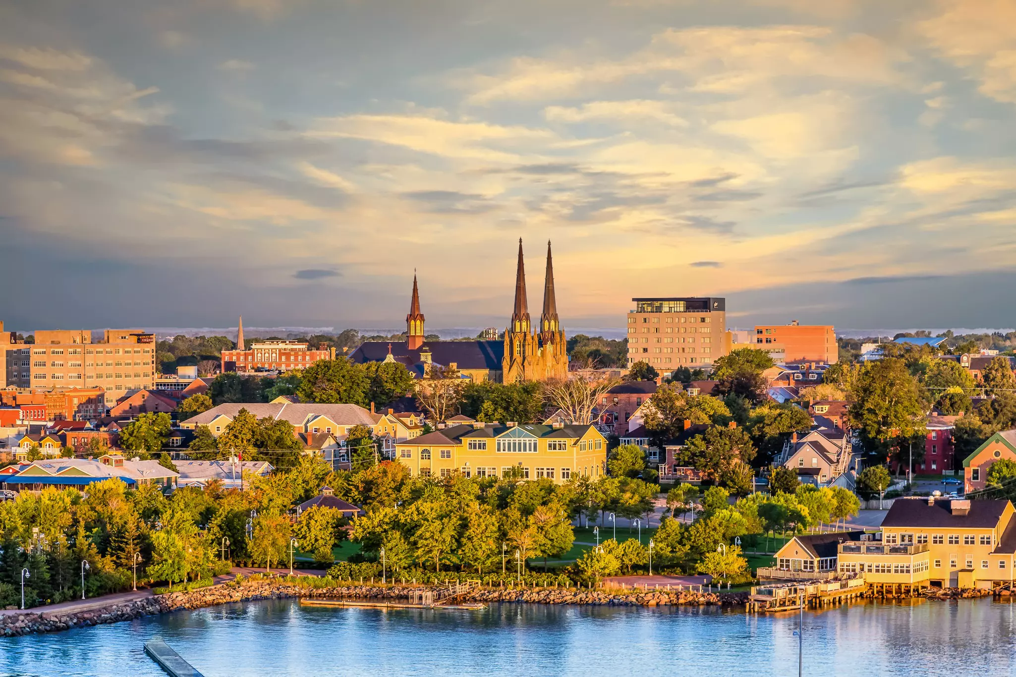 A small waterside city bathed in afternoon light. A two-spired church stands tall above the other buildings.