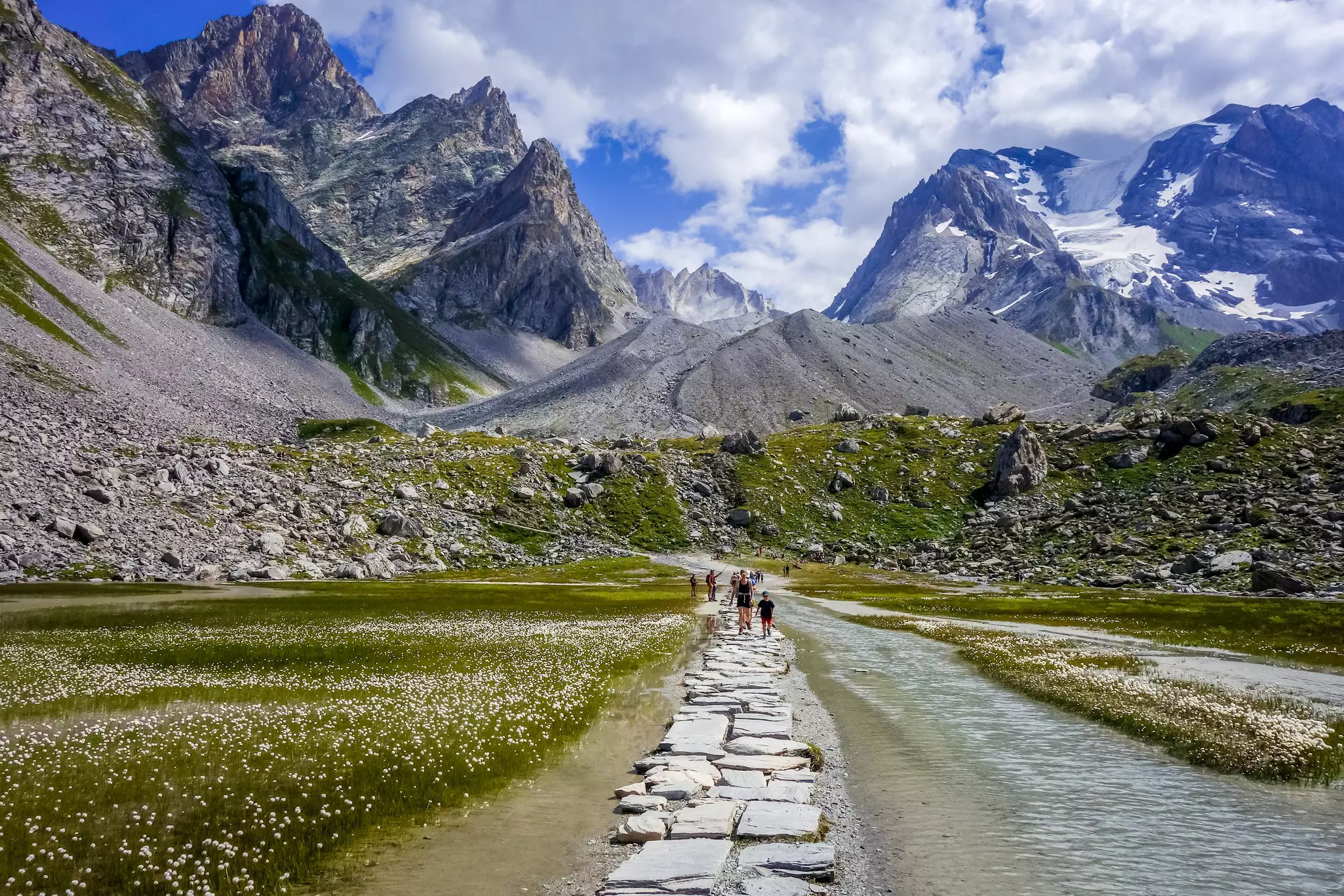 People walk on a stone path through a lakebed in a mountain valley. Rocky peaks hugged by clouds rise sharply from the valley.