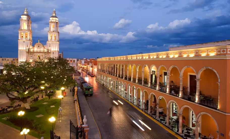 Campeche's twin-towered cathedral lit at dusk beside the main square, with an arched orange colonnade along the street