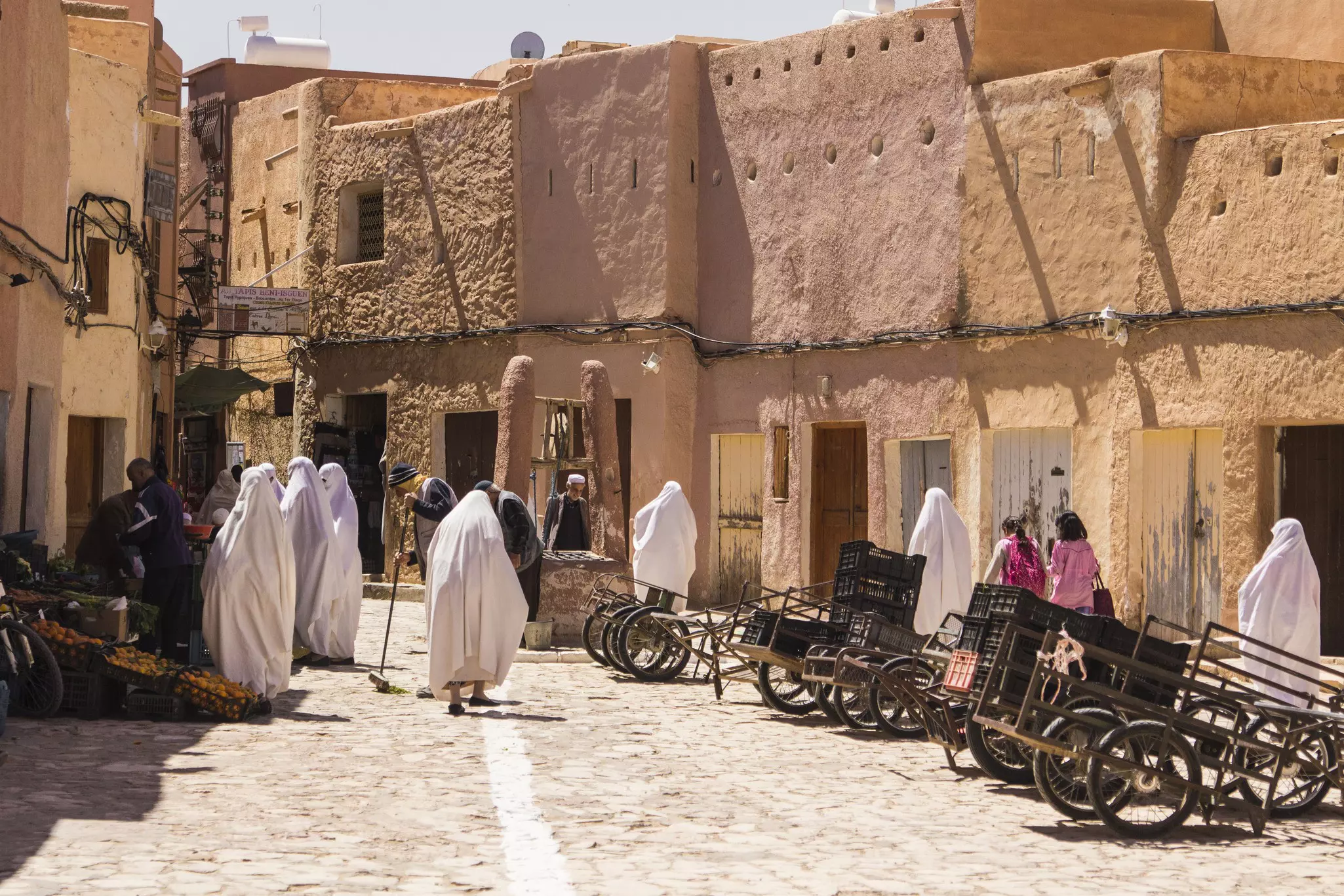 People walking through a market square in a desert town near low-rise stone buildings. Some are covered head to toe in white clothing, others do not have their heads covered.