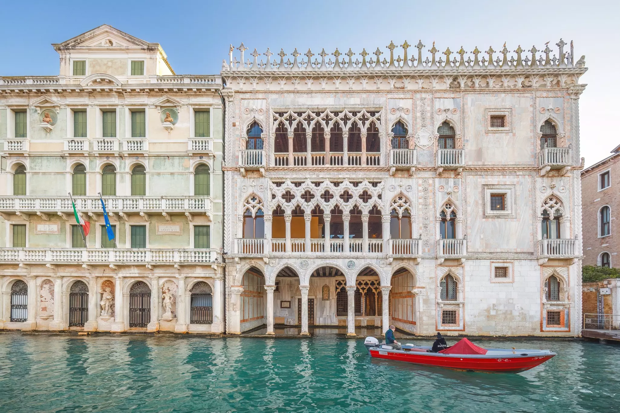 Two men in a small boat alongside a palatial canalside building with Gothic arches over its windows.