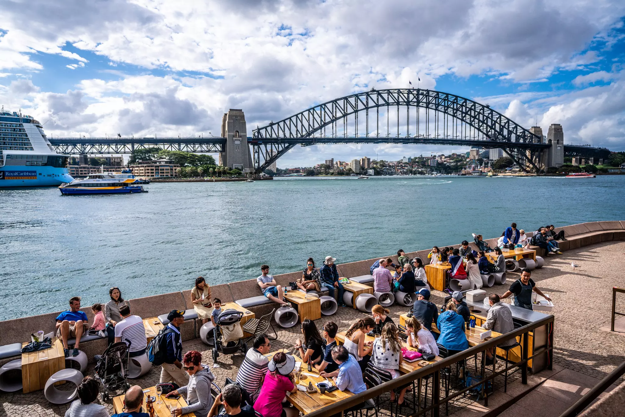 People enjoying a hot summer day at Sydney Opera house outdoor restaurant and bar terrace with Harbour bridge in background.