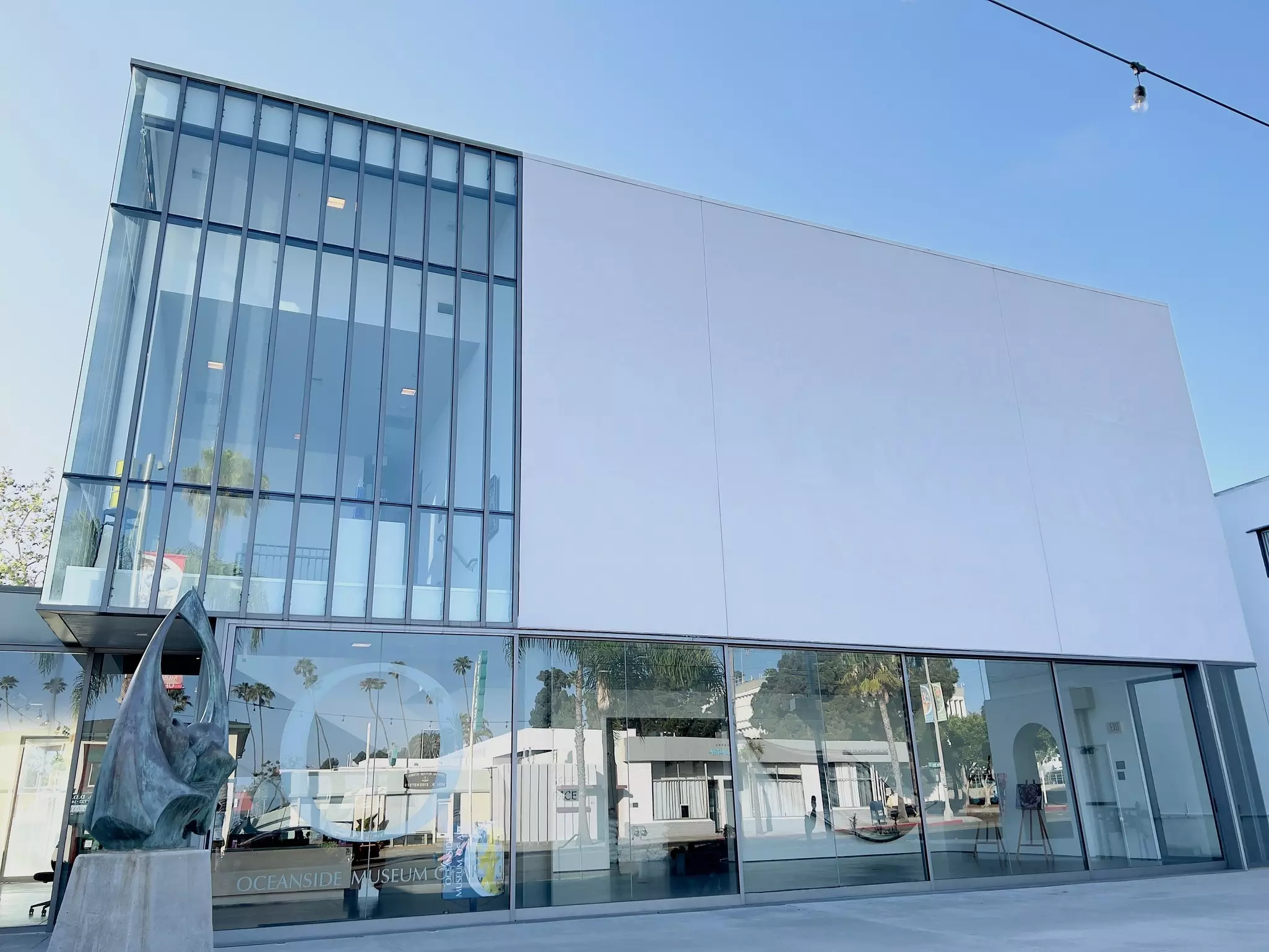 A white building with large windows and a sculpture in front. "Oceanside Museum of Art" is etched in a window.