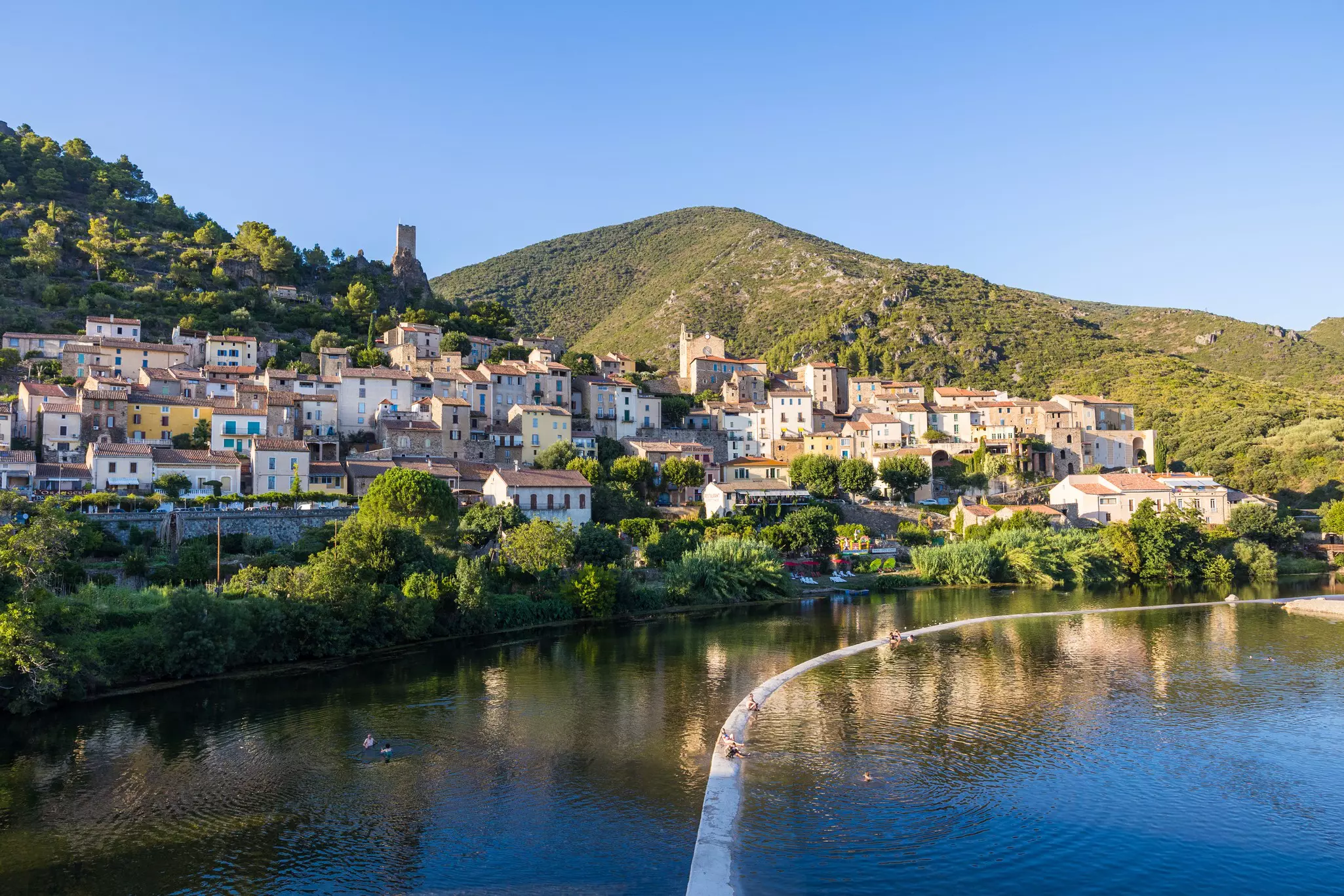 People sit on a weir across a river at the edge of a medieval hilltop village in the evening light.