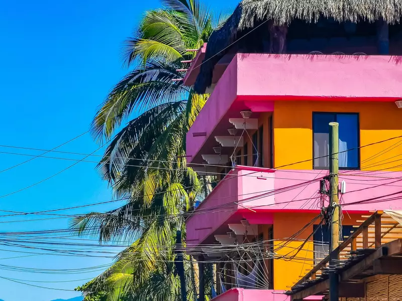 The top stories of a vibrant pink and orange home with balconies and a palm tree in the background on a cloudless, sunny day.