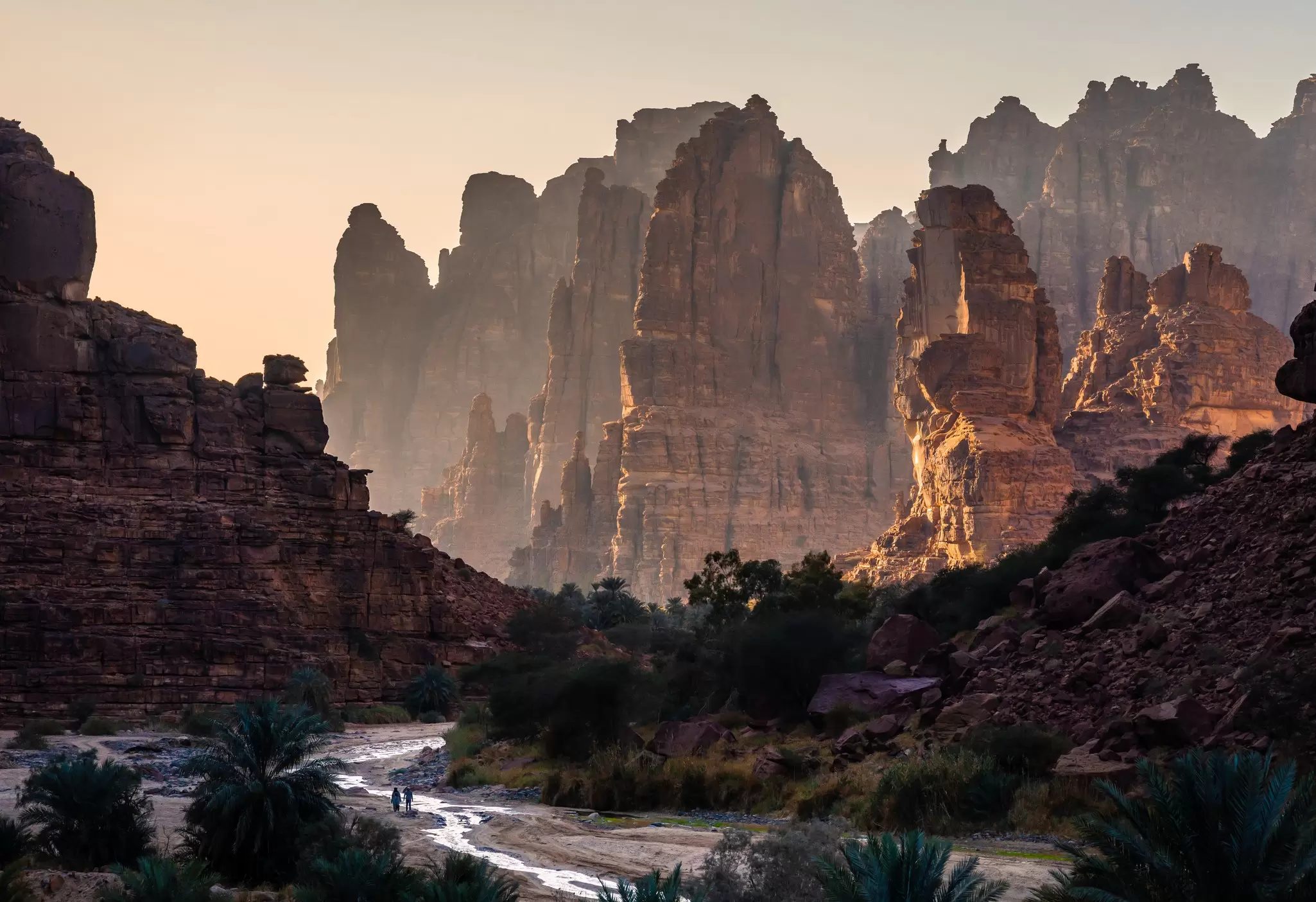Sunset in a rock canyon with two tiny figures by a a small stream.