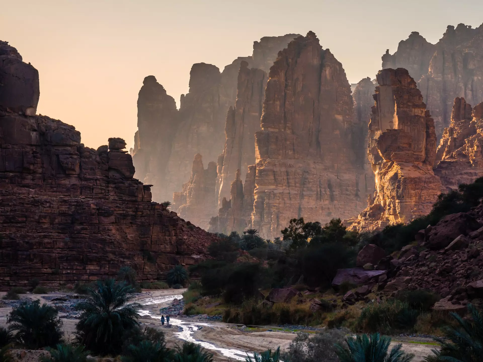Sunset in a rock canyon with two tiny figures by a a small stream.