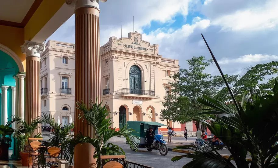 Teatro La Caridad facade in Santa Clara, Cuba, framed by fluted columns and palms of a cafe terrace, a bicitaxi passing