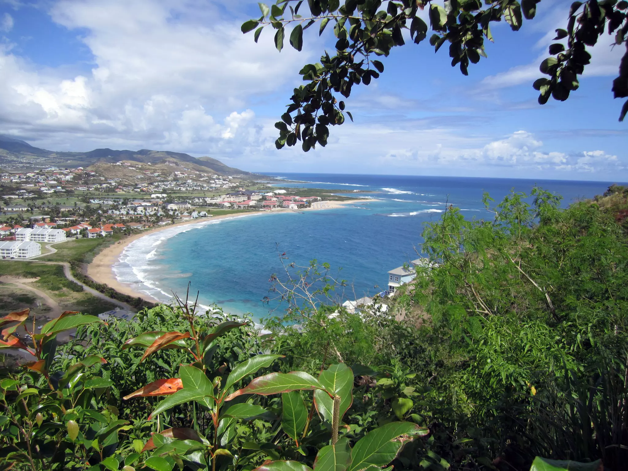 A curve of a sandy bay lapped by the ocean backed by a low-rise town viewed from a hilltop.