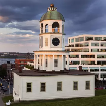 A pinkish glow of sunset illuminates the white siding on a clock tower at the foot of a grassy green hill