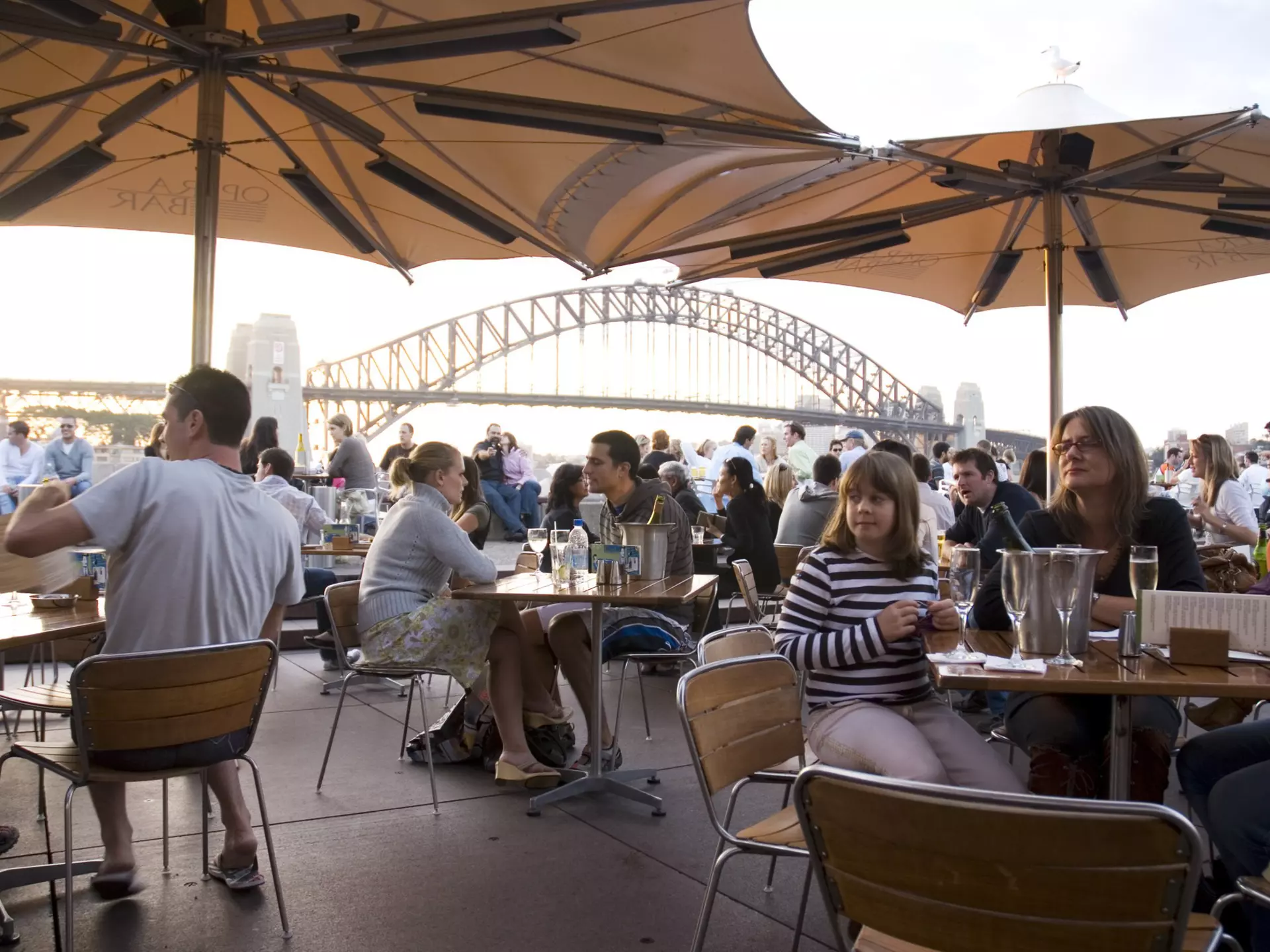People sitting at an outdoor cafe under umbrellas with a view of the Sydney Harbour Bridge.