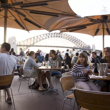 People sitting at an outdoor cafe under umbrellas with a view of the Sydney Harbour Bridge.