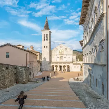 Wide brick steps leading down to a pale stone cathedral with a rose window and tall bell tower, a girl running in front.