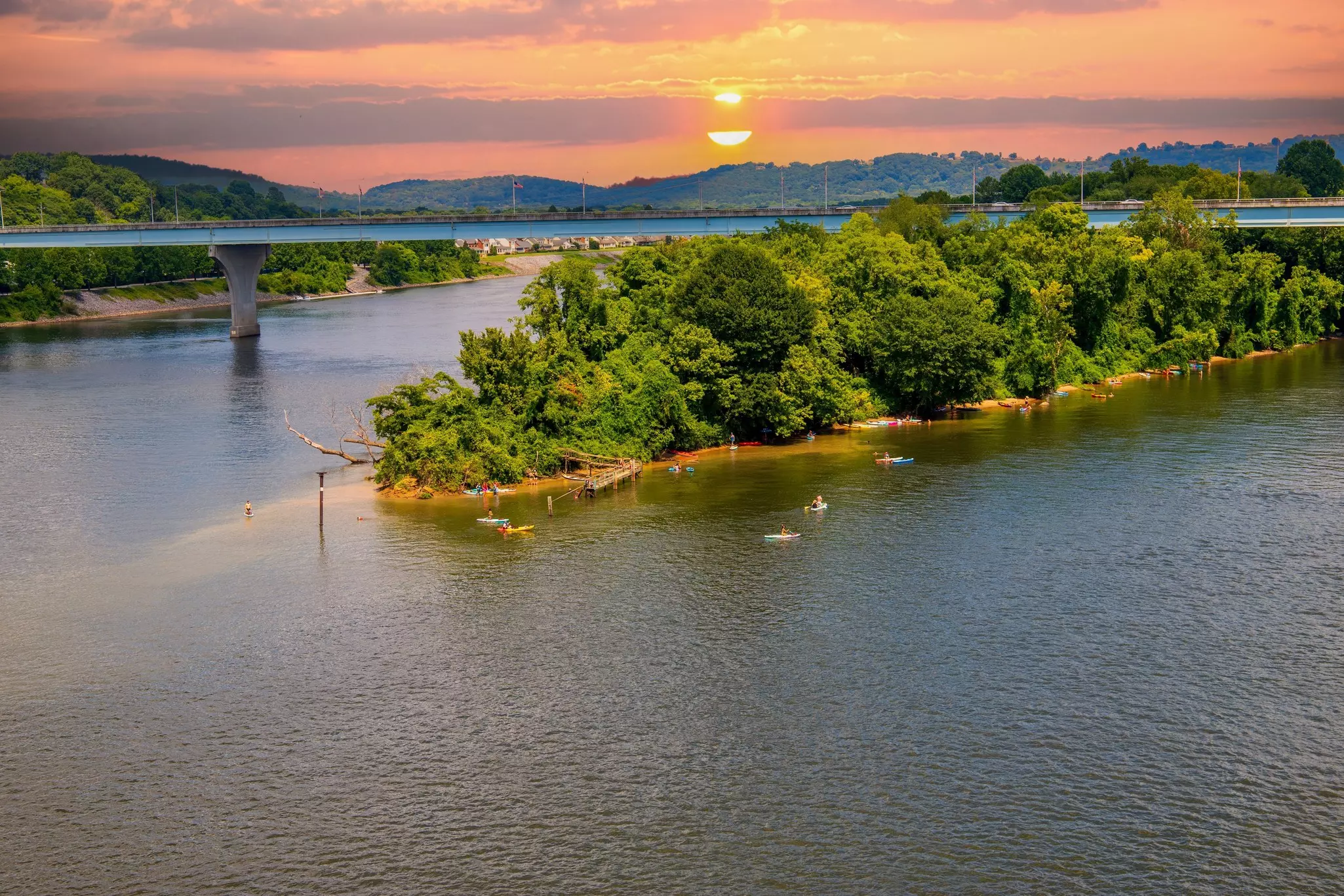 Kayakers on a river near a lush island as the sun sets lighting the sky in orange