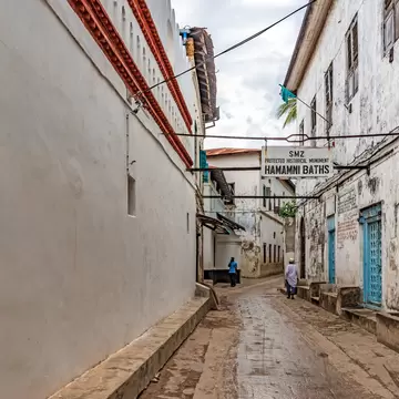 Narrow whitewashed lane in Stone Town with a sign for the Hamamni Baths hanging across it and carved wooden doors