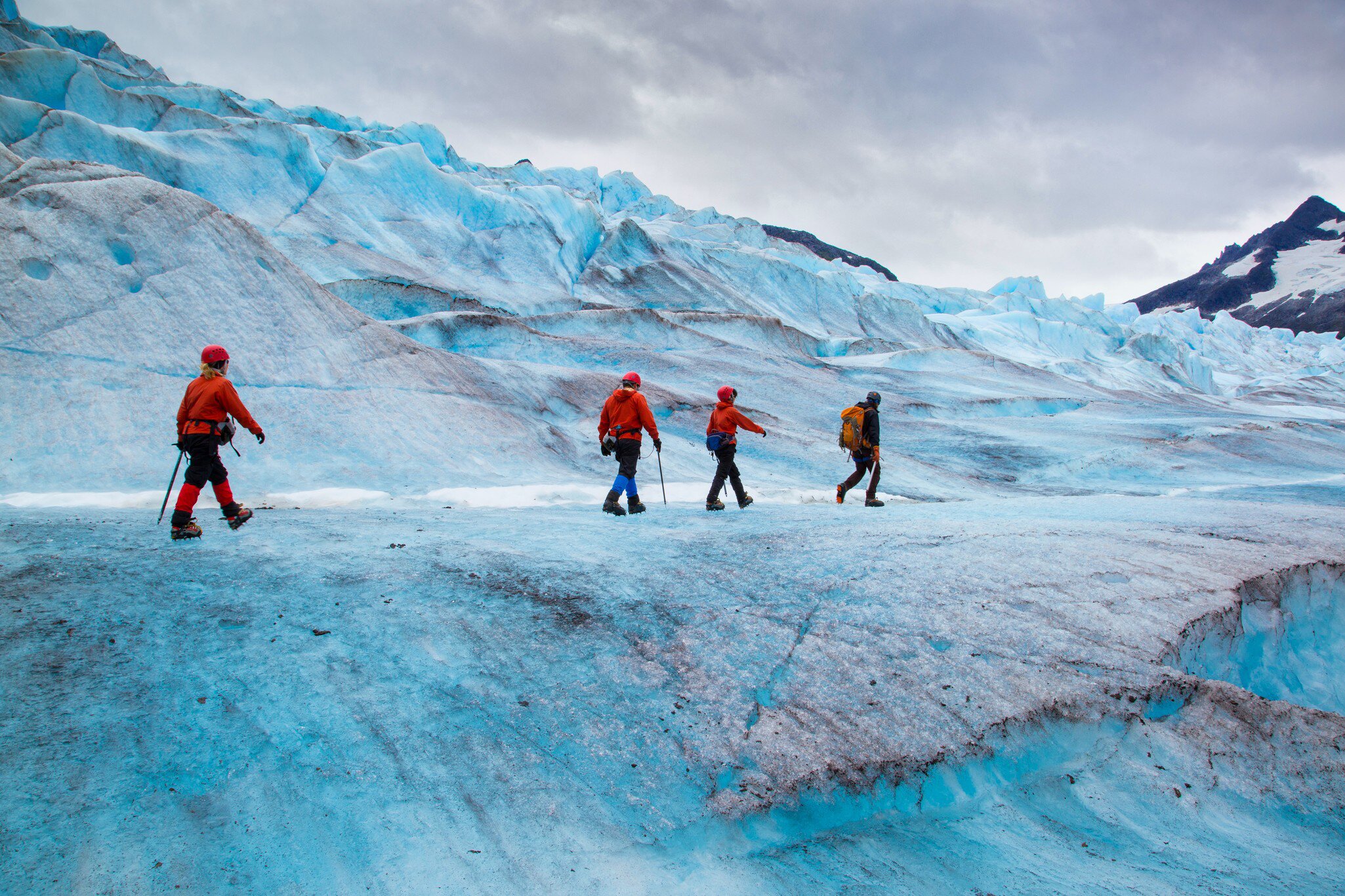Hikers in red jackets and helmets cross rippled blue ice in crampons, a crevasse and grey cloud behind them.