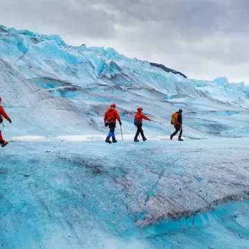 Hikers in red jackets and helmets cross rippled blue ice in crampons, a crevasse and grey cloud behind them.
