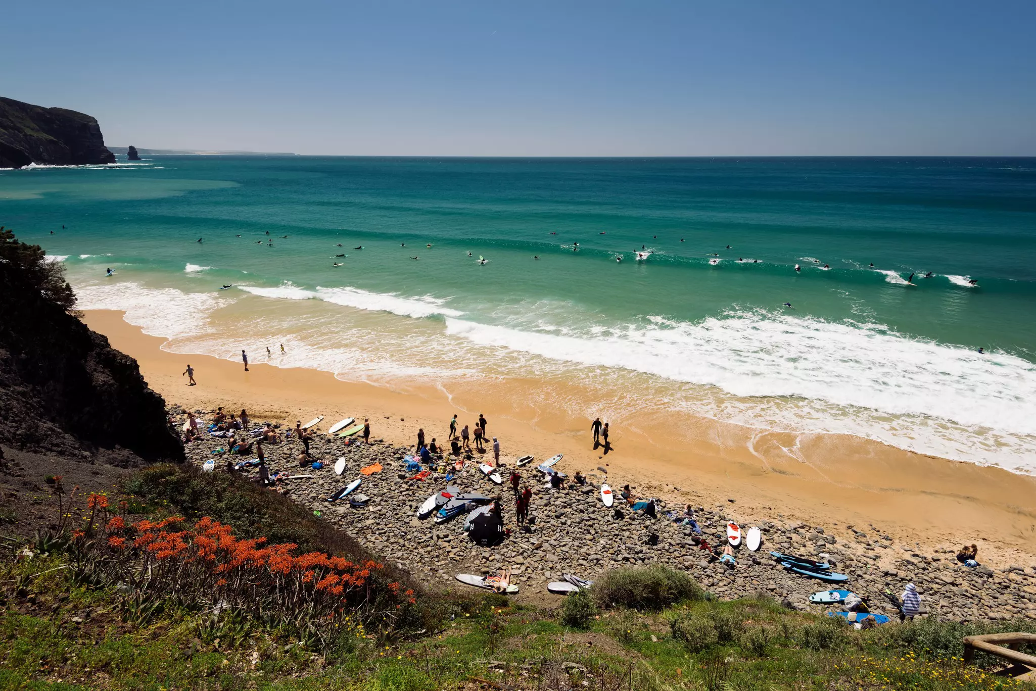 A beach of rocks and sand with surfers gathered on shore and on their boards in the ocean.
