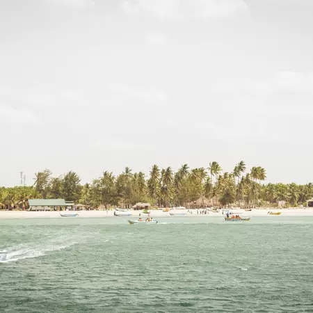 Boats in pale blue water leaving a beach on the shore with sparse greenery.
